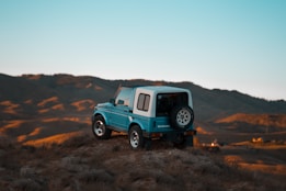 A small off-road vehicle is perched atop a grassy hill, with expansive mountain ranges in the background under a clear blue sky. The vehicle is painted in a vibrant blue with a white roof and appears rugged, suggesting it's suited for adventure.