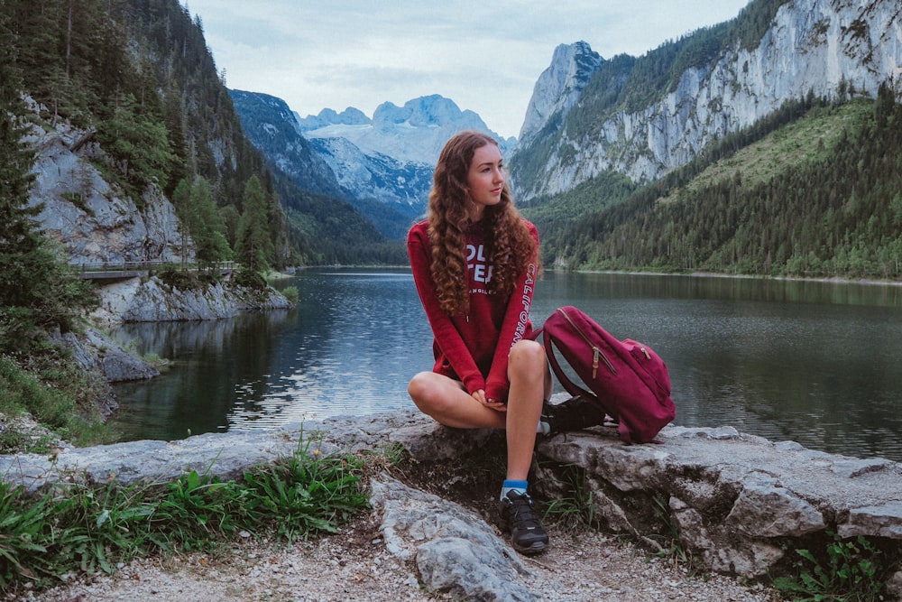 woman in red long sleeve shirt and pink pants sitting on rock near lake during daytime