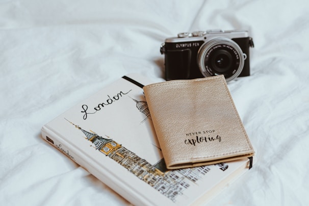 A notebook with an illustration of Big Ben and the word 'London' lies open on a white surface. A vintage-style camera sits adjacent to the notebook, angled slightly towards it. On top of the notebook is a beige passport holder with the words 'Never Stop Exploring' printed on it.