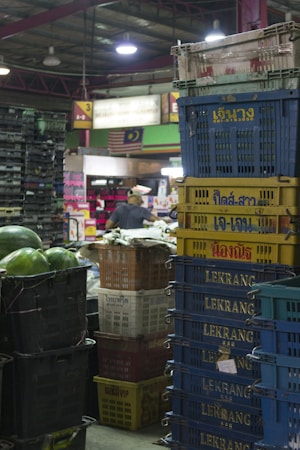 A market scene with large stacks of plastic crates in various colors, some with writing on them. Watermelons can be seen in black crates on the left, and a person wearing a cap is in the background among more crates and market goods. There are signs, lighting, and a mix of green, red, and white colors throughout the space.