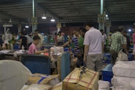 A bustling indoor market scene with many people engaged in buying and selling activities. There are stacks of packaged goods and containers around, and vendors are interacting with customers. The structure suggests a large warehouse with numbered sections on the pillars.