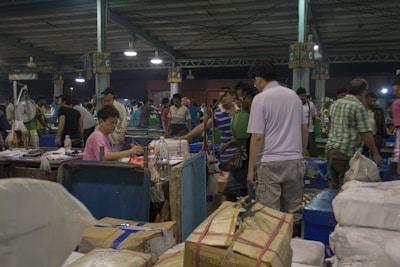 A bustling indoor market scene with many people engaged in buying and selling activities. There are stacks of packaged goods and containers around, and vendors are interacting with customers. The structure suggests a large warehouse with numbered sections on the pillars.