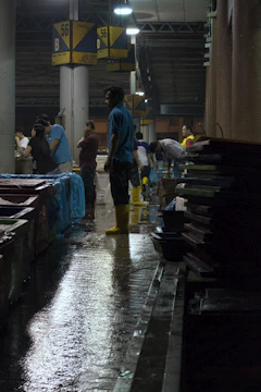 Several people are working in a dimly lit indoor market or warehouse. The floor is wet, and some individuals are wearing yellow boots, indicating it might be a fish or seafood market. Overhead lights cast reflections on the wet ground, and signs with the number 56 and the letter B are visible, possibly marking sections.