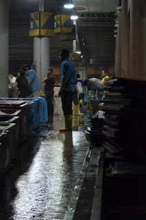 Several people are working in a dimly lit indoor market or warehouse. The floor is wet, and some individuals are wearing yellow boots, indicating it might be a fish or seafood market. Overhead lights cast reflections on the wet ground, and signs with the number 56 and the letter B are visible, possibly marking sections.