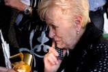 An elderly woman with short blonde hair is holding a phone to her ear. She is wearing a black and white patterned outfit and pearl earrings. In front of her, there is a wrapped gift with shiny gold and red ribbon. Other people are partly visible in the background.