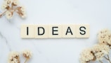 Five ivory-colored Scrabble tiles arranged to spell the word 'IDEAS' on a white marble surface. Surrounding the tiles are delicate, dried flowers, adding an elegant and creative touch to the composition.
