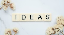 Five ivory-colored Scrabble tiles arranged to spell the word 'IDEAS' on a white marble surface. Surrounding the tiles are delicate, dried flowers, adding an elegant and creative touch to the composition.