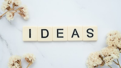 Five ivory-colored Scrabble tiles arranged to spell the word 'IDEAS' on a white marble surface. Surrounding the tiles are delicate, dried flowers, adding an elegant and creative touch to the composition.