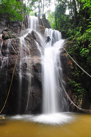 A cascading waterfall flows down a rocky cliff surrounded by lush green foliage. A person is seen descending on a rope in what appears to be a rappelling activity. The water is frothy as it hits the pool below.