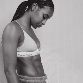 A person with braided hair is seen in profile wearing a Calvin Klein sports bra and sweatpants. The image is in black and white, highlighting the person's toned physique and contemplative posture against a plain background.