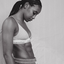A person with braided hair is seen in profile wearing a Calvin Klein sports bra and sweatpants. The image is in black and white, highlighting the person's toned physique and contemplative posture against a plain background.