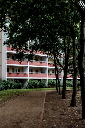 A pathway lined with trees leading towards a residential building with red and white balconies. The building has multiple stories, and each balcony features potted plants. The area is shaded by dense tree foliage, creating a serene and calm atmosphere.