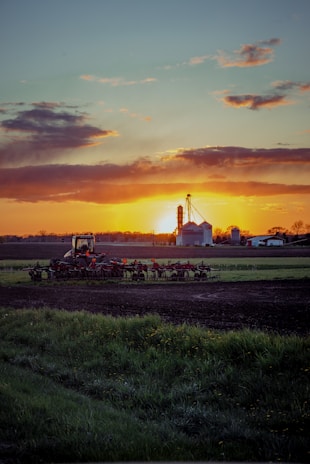 In a rural landscape at sunset, there is a tractor with farming equipment in the foreground, partially surrounded by green grass and soil. In the background, there are silos and farm buildings under a cloudy sky, illuminated by the setting sun.