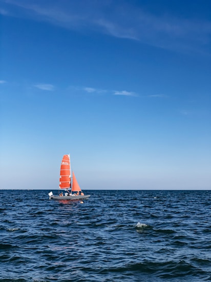 A sailboat with bright orange sails glides across a vast, calm sea under a clear blue sky. The water is slightly rippled, reflecting the sunlight and creating a serene atmosphere.