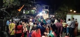 A lively street scene at night features a large group of people gathered, many dressed in colorful attire. In the background, a vehicle adorned with lights, resembling a festive or ceremonial float, is visible along with surrounding houses. A sense of community and celebration is evident, with some people holding candles or small lights.
