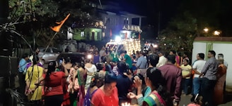 A lively street scene at night features a large group of people gathered, many dressed in colorful attire. In the background, a vehicle adorned with lights, resembling a festive or ceremonial float, is visible along with surrounding houses. A sense of community and celebration is evident, with some people holding candles or small lights.