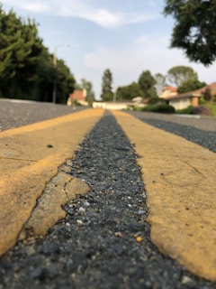 A close-up view of a textured asphalt road with two yellow lines running down the center. The road is surrounded by greenery and residential buildings in the background, with trees lining both sides. The perspective is low and focused on the road, creating a sense of depth.