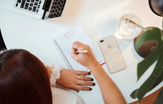 A person is writing in a spiral notebook on a white desk. Nearby, there is a laptop partially visible, a smartphone lying face up, a glass vase, and a green plant. The person is wearing a white shirt and has black nail polish.