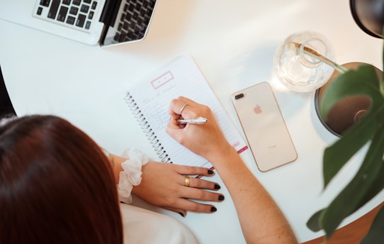 A person is writing in a spiral notebook on a white desk. Nearby, there is a laptop partially visible, a smartphone lying face up, a glass vase, and a green plant. The person is wearing a white shirt and has black nail polish.