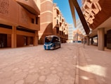 A modern architectural street scene featuring a futuristic driverless vehicle. The buildings have an innovative design with intricate patterns and rounded shapes, reflecting Middle Eastern architecture. The scene is well-lit with clear blue skies above.