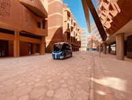 A modern architectural street scene featuring a futuristic driverless vehicle. The buildings have an innovative design with intricate patterns and rounded shapes, reflecting Middle Eastern architecture. The scene is well-lit with clear blue skies above.