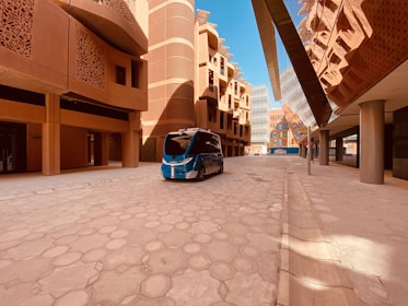 A modern architectural street scene featuring a futuristic driverless vehicle. The buildings have an innovative design with intricate patterns and rounded shapes, reflecting Middle Eastern architecture. The scene is well-lit with clear blue skies above.