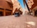 A modern architectural street scene featuring a futuristic driverless vehicle. The buildings have an innovative design with intricate patterns and rounded shapes, reflecting Middle Eastern architecture. The scene is well-lit with clear blue skies above.