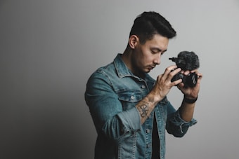 A person wearing a denim jacket is holding a camera with a microphone attachment. The individual appears focused on the camera while standing against a plain background. A tattoo is visible on the person's forearm.