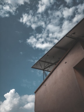 The image contains a view of a building with a modern architectural design, featuring a canopy or awning. In the background, there is a partly cloudy sky, with soft, white clouds scattered across a blue sky.