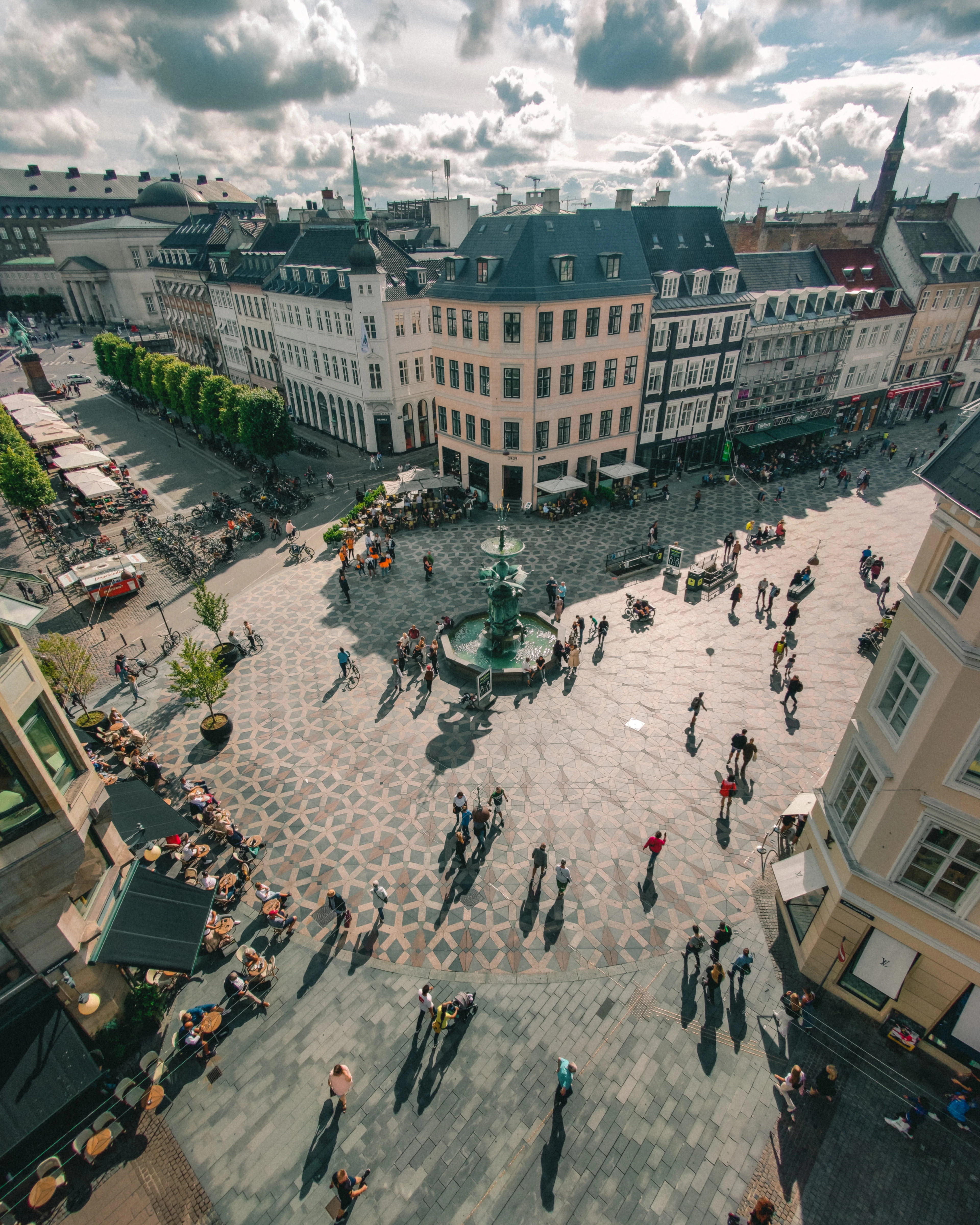 Aerial view of a paved city square with a central fountain, long shadows, and people crossing under a cloud-streaked sky