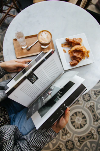 A person reading a book beside a coffee and croissants on a table