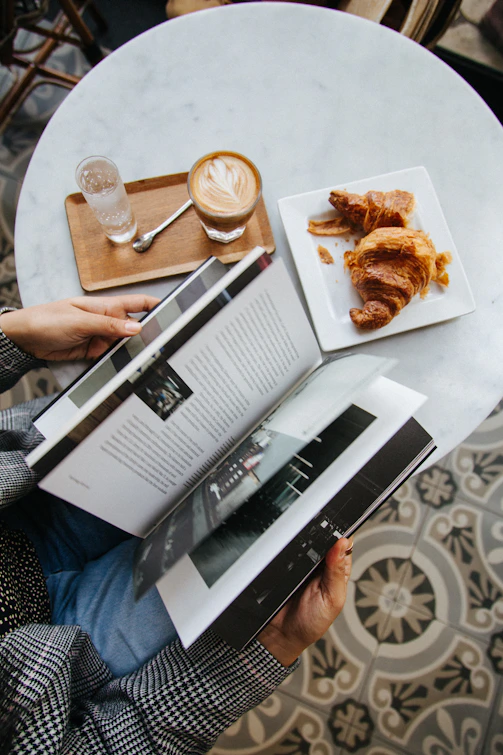 A person reading a book beside a coffee and croissants on a table