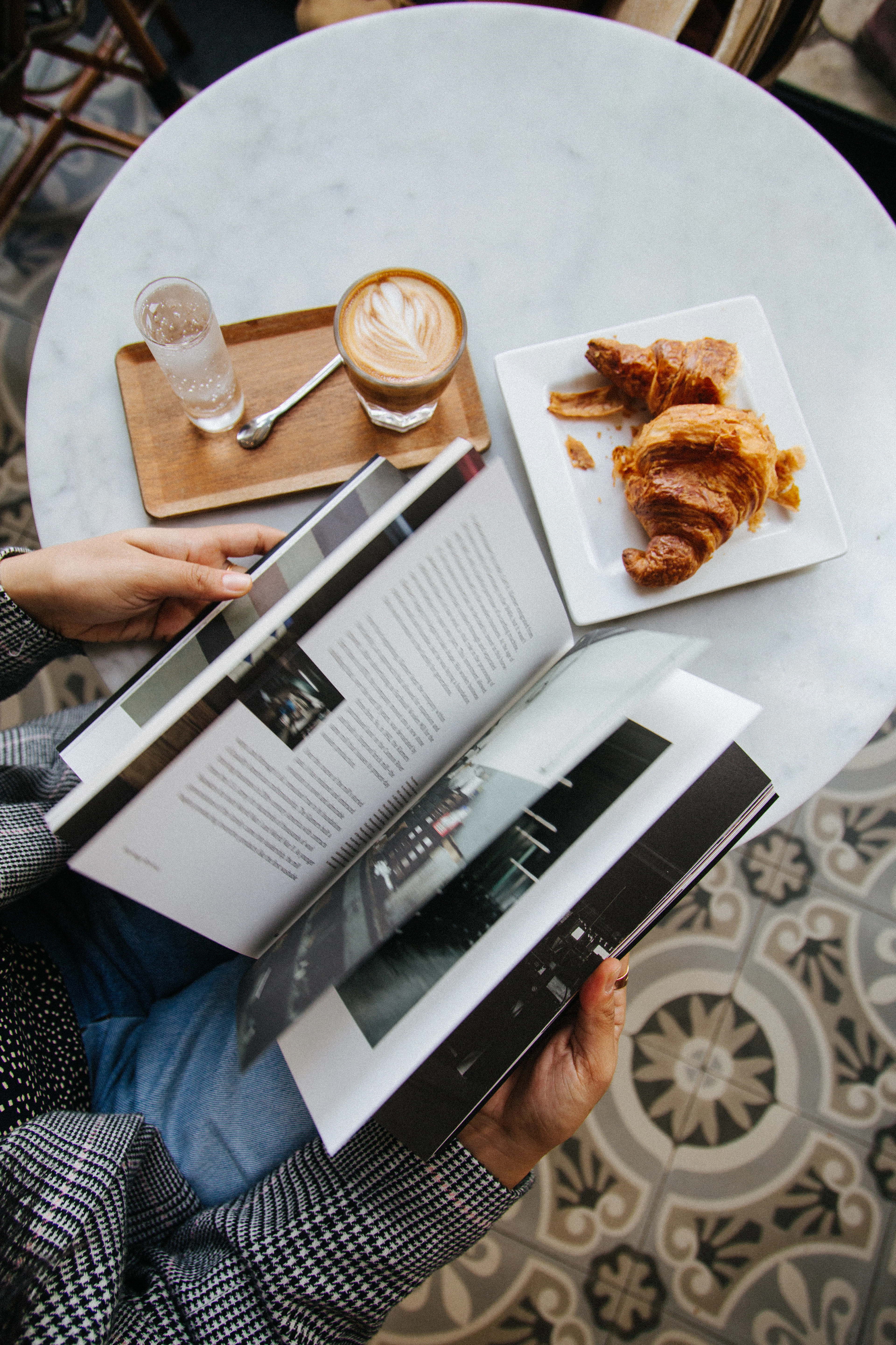 A marble café table in Paris set with croissants, a latte and an open book above a patterned tile floor