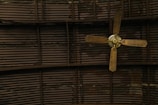 A ceiling fan with four wooden blades is mounted against a corrugated metal ceiling. The blades appear slightly worn and the metal ceiling has a dark, industrial look with visible lines and sections.