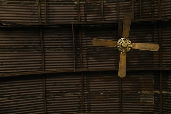 A ceiling fan with four wooden blades is mounted against a corrugated metal ceiling. The blades appear slightly worn and the metal ceiling has a dark, industrial look with visible lines and sections.