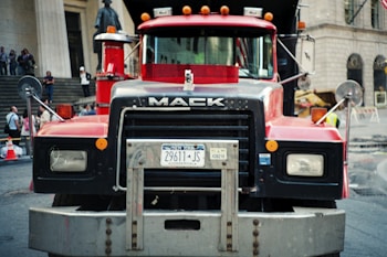 A large red Mack truck is positioned prominently in the center, with its grille and front details clearly visible. The truck features a New York license plate, and various elements like mirrors and lights on the exterior. In the background, a busy urban setting shows people standing on stairs, an entrance to a building, and some street cones, suggesting a construction or city environment.