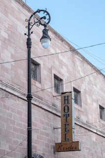 A vintage street lamp stands in front of a light pink brick building with a vertical sign reading 'HOTEL Rosales' in bold black letters. The building features several small windows with decorative bars, and numerous wires are visible against a clear blue sky.