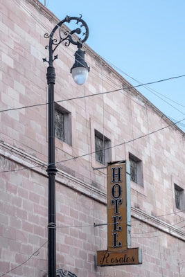 A vintage street lamp stands in front of a light pink brick building with a vertical sign reading 'HOTEL Rosales' in bold black letters. The building features several small windows with decorative bars, and numerous wires are visible against a clear blue sky.