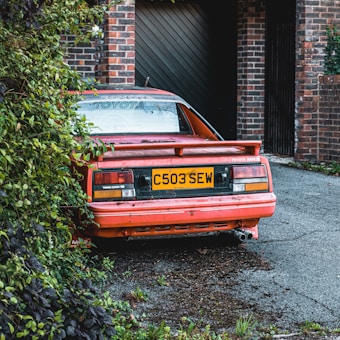 A vintage red Toyota car with a visible rear license plate, partially hidden by foliage and parked near a brick garage. The car appears weathered, with some dirt and vegetation covering parts of it.