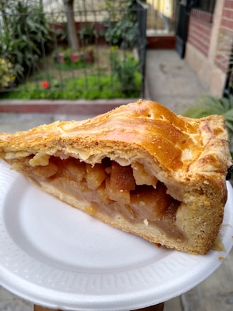 A slice of apple pie with a golden-brown crust served on a white disposable plate. The filling is visible, displaying chunks of cooked apples. In the background, there is a garden with green foliage and a hint of red flowers, alongside the exterior walls of a building.
