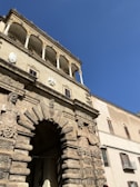 An ornate stone archway adorned with intricate carvings and sculptures of human figures, embedded in a historic building facade. Above the archway, a series of elegant columns support a decorative balcony. The architecture exhibits classical design elements with detailed stonework. The clear blue sky provides a contrasting backdrop to the Sandy cream-colored structure.