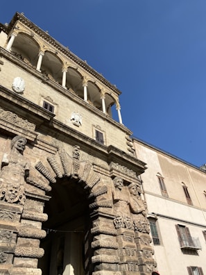 An ornate stone archway adorned with intricate carvings and sculptures of human figures, embedded in a historic building facade. Above the archway, a series of elegant columns support a decorative balcony. The architecture exhibits classical design elements with detailed stonework. The clear blue sky provides a contrasting backdrop to the Sandy cream-colored structure.