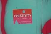 A sticker with a gradient from orange to pink that reads 'Creativity Express Yourself' is pasted on a teal surface. The sticker also features a design with a circle and a triangle, and is surrounded by red graffiti on the left and bottom edges of the image.