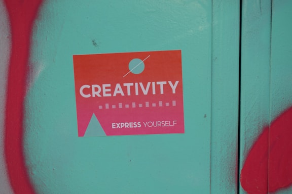 A sticker with a gradient from orange to pink that reads 'Creativity Express Yourself' is pasted on a teal surface. The sticker also features a design with a circle and a triangle, and is surrounded by red graffiti on the left and bottom edges of the image.