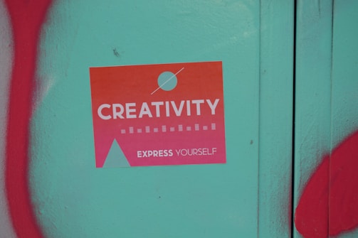 A sticker with a gradient from orange to pink that reads 'Creativity Express Yourself' is pasted on a teal surface. The sticker also features a design with a circle and a triangle, and is surrounded by red graffiti on the left and bottom edges of the image.