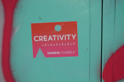 A sticker with a gradient from orange to pink that reads 'Creativity Express Yourself' is pasted on a teal surface. The sticker also features a design with a circle and a triangle, and is surrounded by red graffiti on the left and bottom edges of the image.