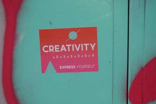 A sticker with a gradient from orange to pink that reads 'Creativity Express Yourself' is pasted on a teal surface. The sticker also features a design with a circle and a triangle, and is surrounded by red graffiti on the left and bottom edges of the image.
