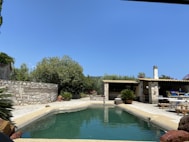 A rectangular swimming pool surrounded by a stone patio is bordered by lush greenery and shrubs. There is a rustic outdoor structure with a wooden roof and stone pillars, which includes a small dining area with tables and chairs. The sky is clear and blue, suggesting a calm and sunny day.