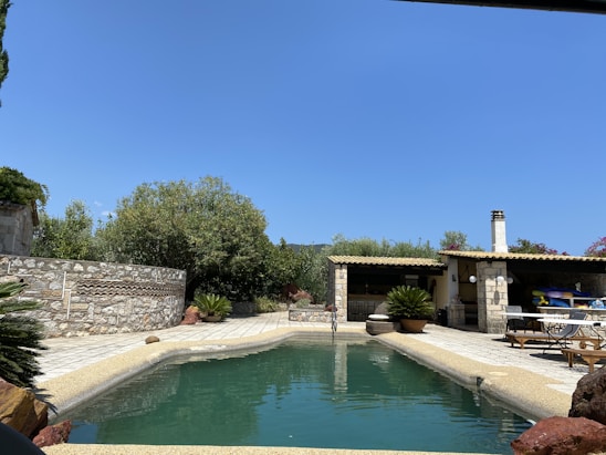 A rectangular swimming pool surrounded by a stone patio is bordered by lush greenery and shrubs. There is a rustic outdoor structure with a wooden roof and stone pillars, which includes a small dining area with tables and chairs. The sky is clear and blue, suggesting a calm and sunny day.