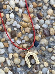 A red string necklace with a horseshoe-shaped pendant inscribed with 'Soulmate Magnet' rests on a surface covered with small, multicolored pebbles.